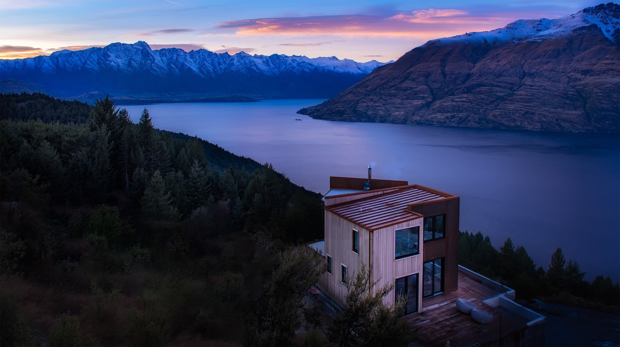 Luc 22 at dusk, a cedar and corten-steel house on a forested ridge above Lake Wakatipu