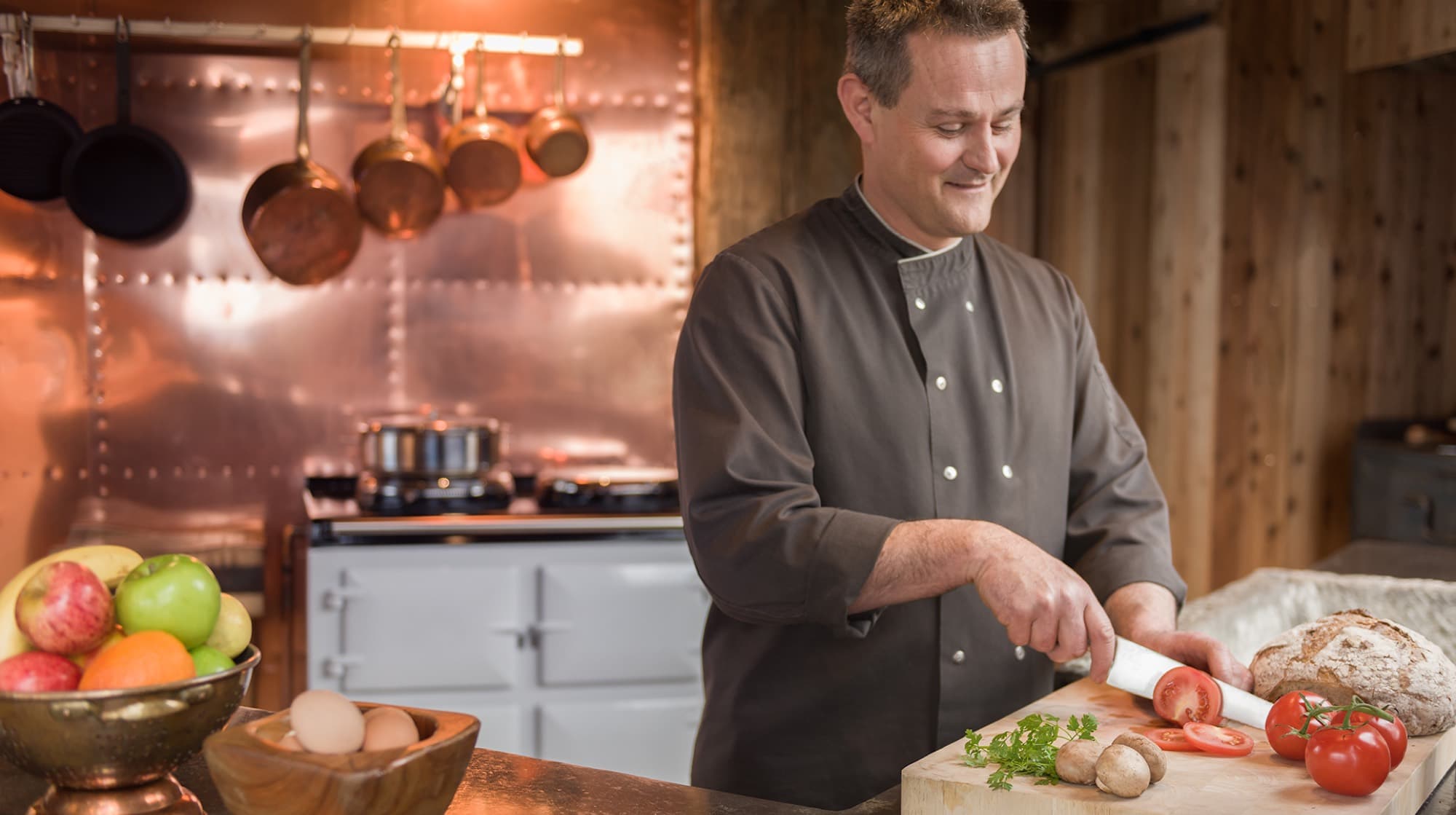 Luc slicing vegetables at the bench of the copper-clad kitchen, an AGA range and hanging copper pans behind him