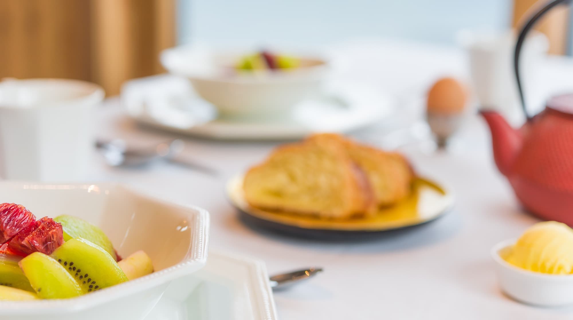 Breakfast laid on white linen with fresh fruit, warm pastries and a red cast-iron teapot
