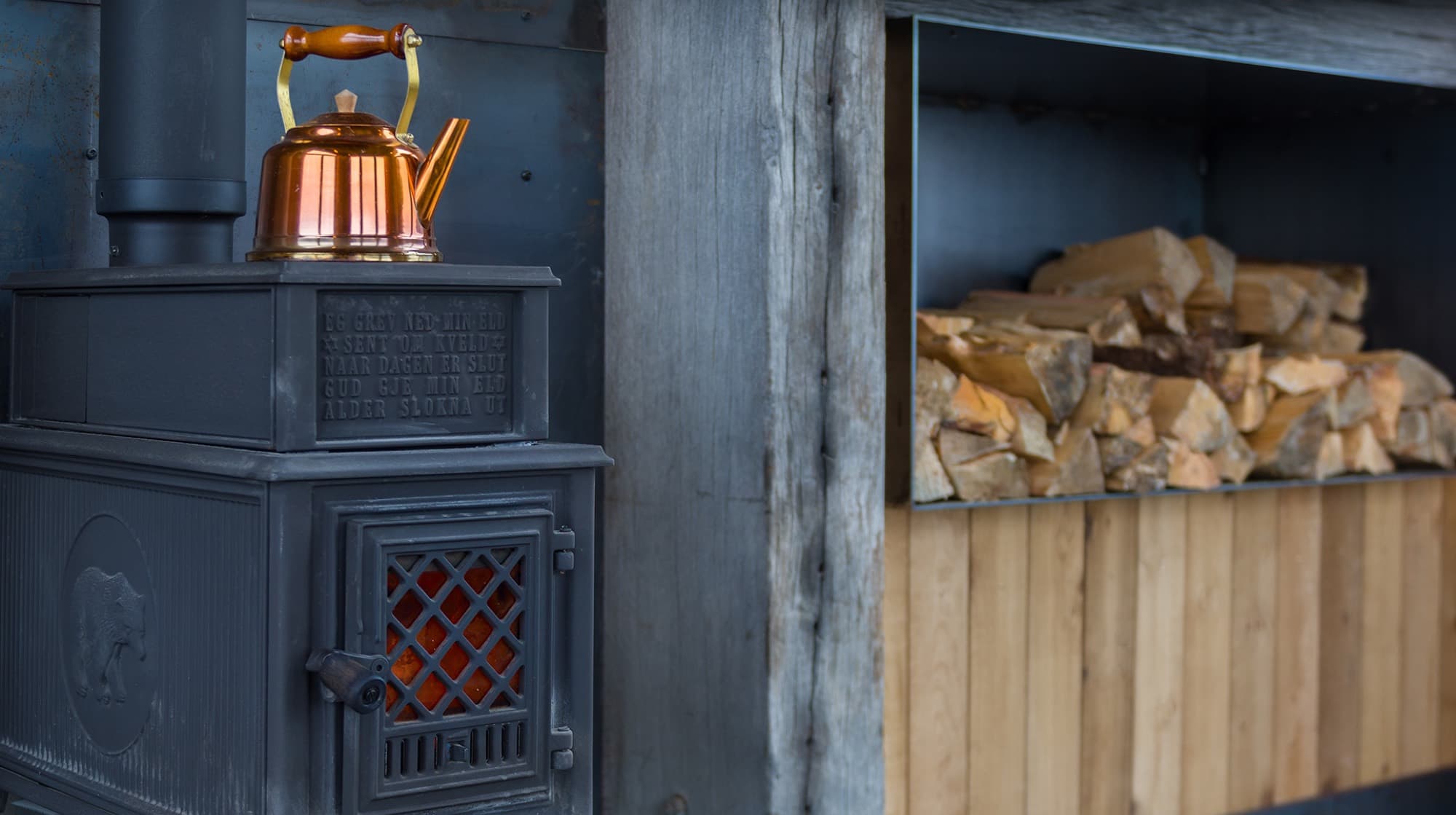 The Jotul woodburner alight with a copper kettle on top and a stacked woodstore beside it