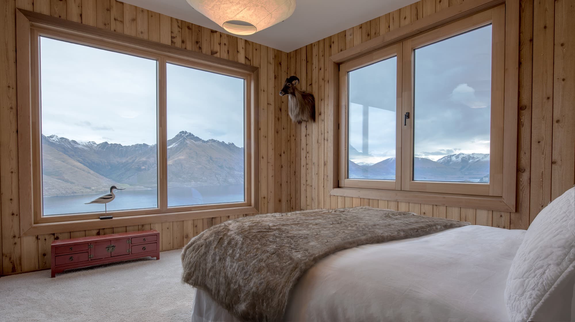 The master suite, a bed under fur throws in a cedar-lined room with two windows onto the lake and mountains