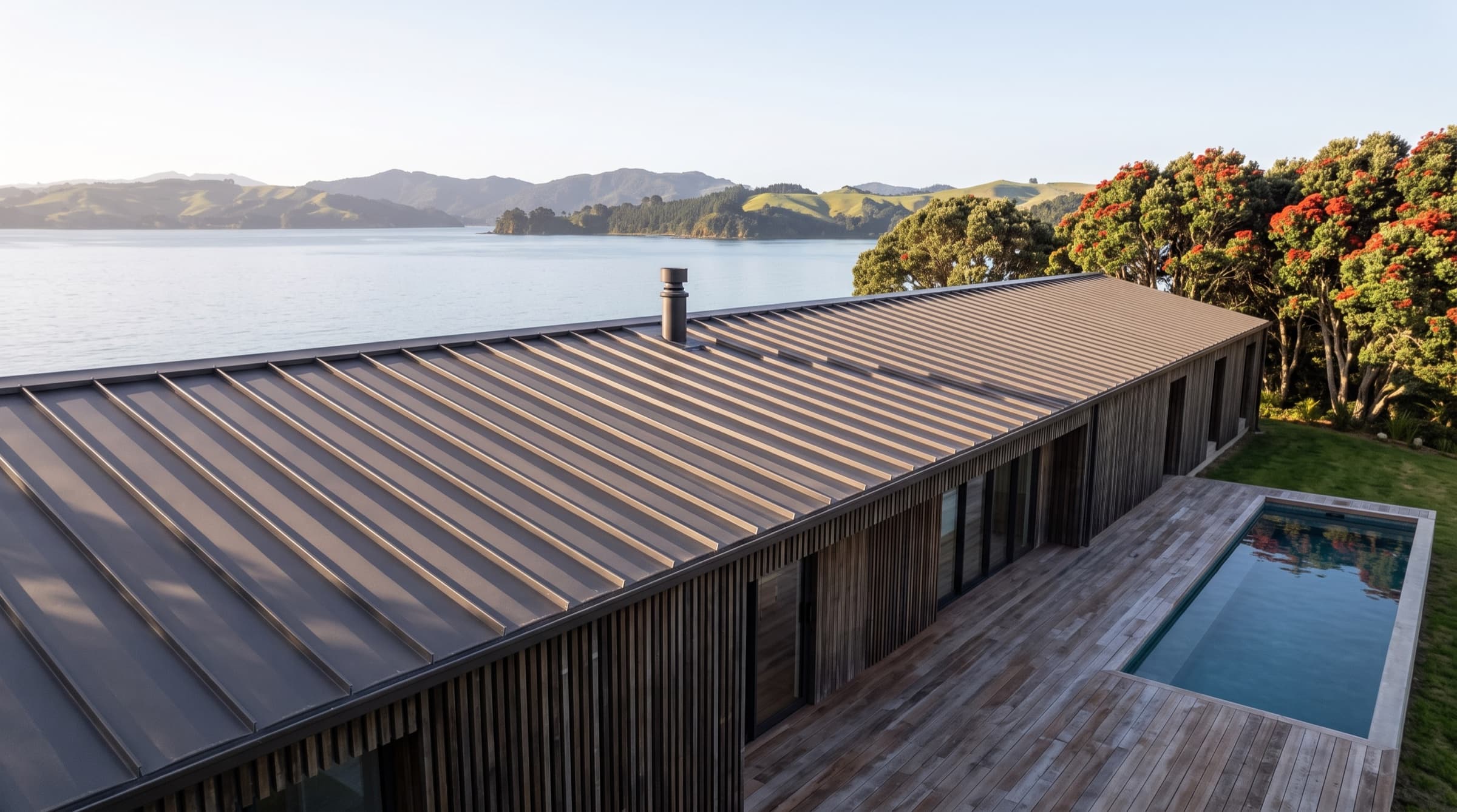 The long low standing-seam roofline of Tideline House from above, the bay filling the distance